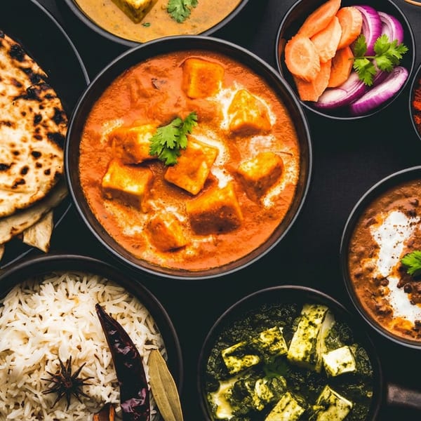Overhead view of various Indian dishes: naan, rice, paneer curries, dal, and spices on a dark table.