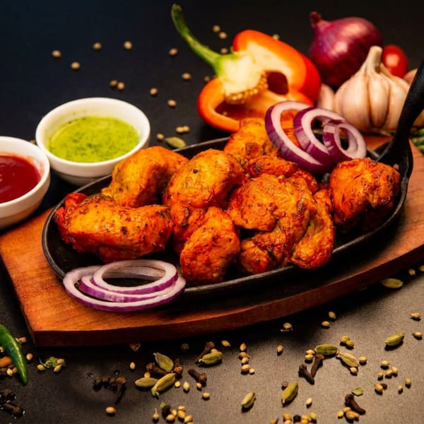 Grilled chicken pieces in a pan with sauces, red onion, garlic, and scattered spices on a dark surface.