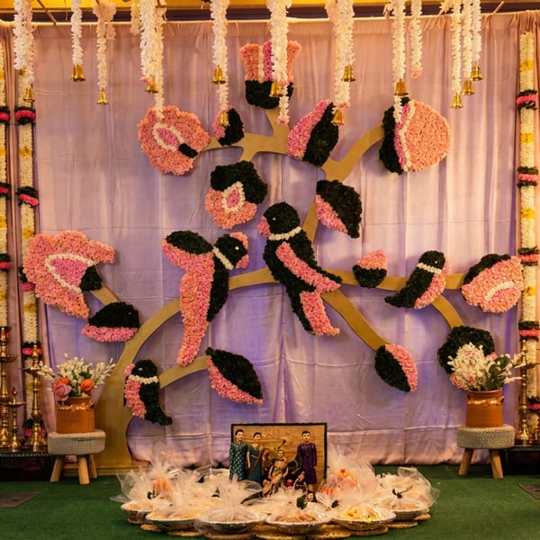 Elaborate floral stage decoration with pink and black birds, garlands, traditional lamps, and offerings.